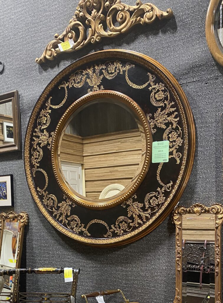 Large round vintage mirror with an ornate black and gold frame and intricate gold floral patterns, displayed among other secondhand mirrors and unique salvaged decor at Second Chance, a Baltimore non-profit.